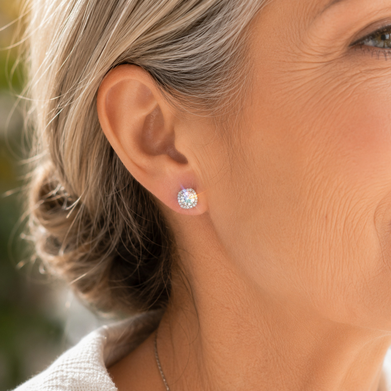 Close-up of a mature woman wearing 925 sterling silver cushion halo moissanite stud earrings on her right ear in natural sunlight, showcasing brilliant sparkle, rainbow fire, and timeless elegance.