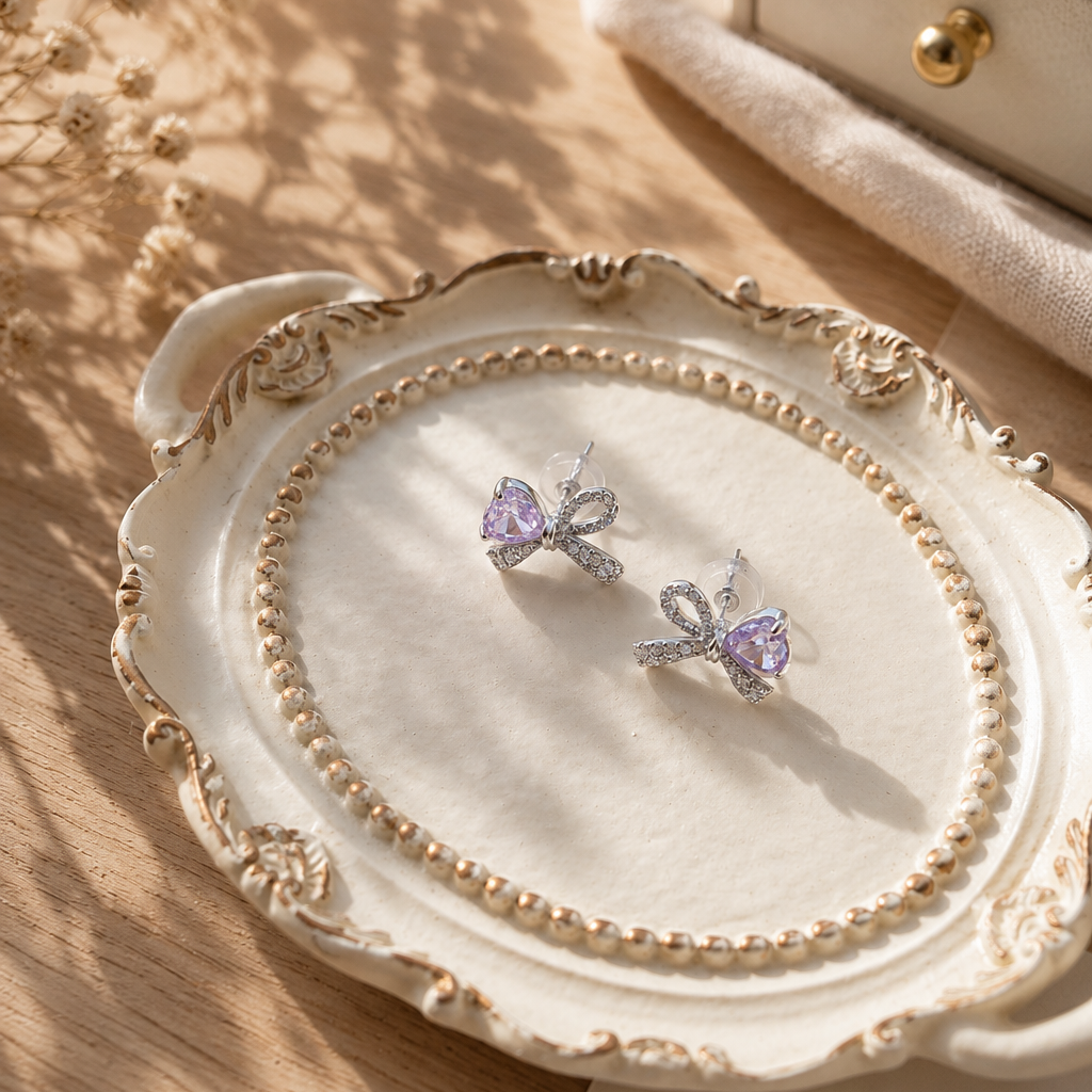 Flat lay of 925 sterling silver bow stud earrings featuring sparkling Snowflake Cut purple heart cubic zirconia on a vintage ceramic tray, styled with warm natural sunlight for an elegant handmade jewelry presentation.