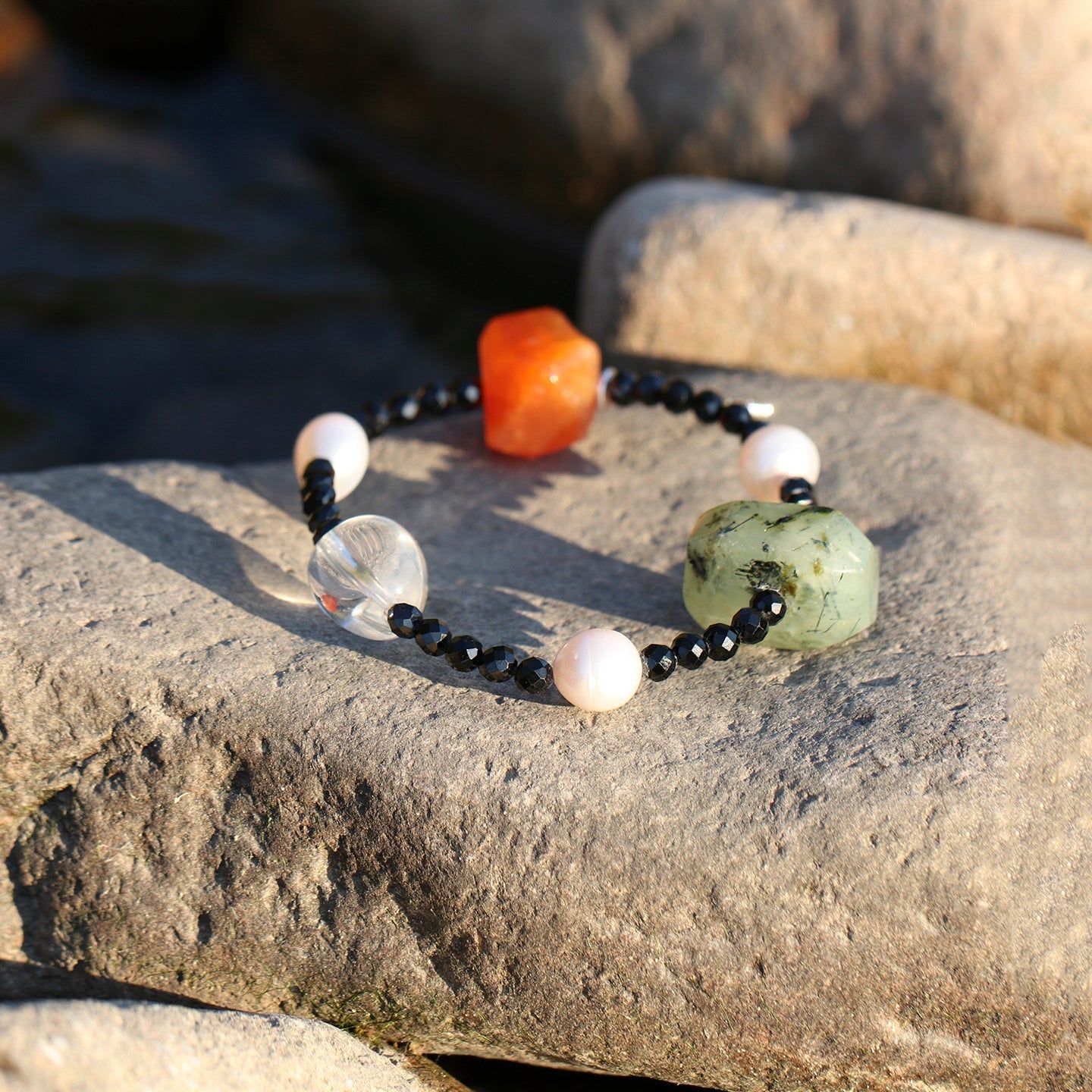 Handcrafted natural carnelian, prehnite, clear quartz, black onyx and freshwater pearl bracelet displayed on sunlit natural stone, showcasing organic textures and timeless artisan craftsmanship.