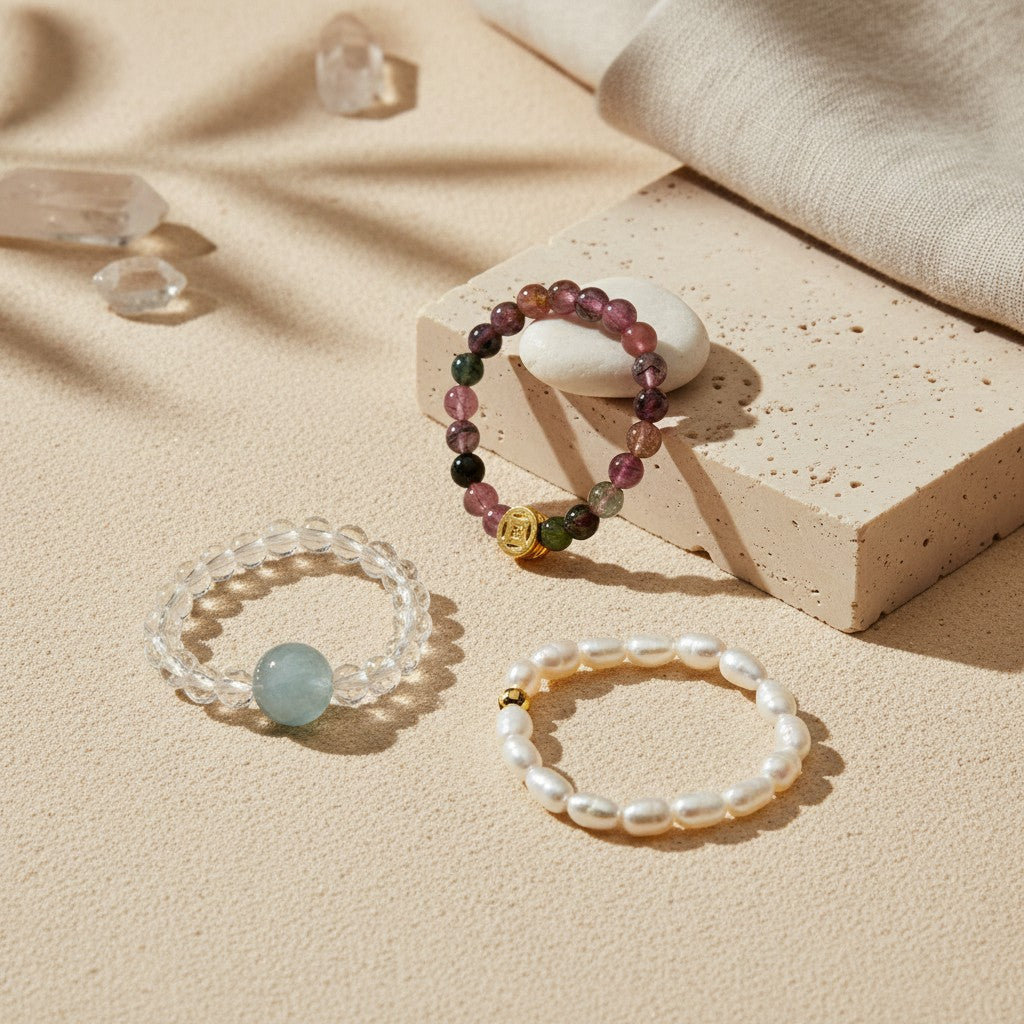 Natural gemstone stretch ring collection featuring Tourmaline, Aquamarine, and Freshwater Pearl designs displayed on a neutral stone platform with soft natural sunlight.