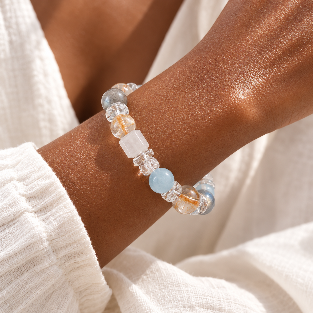 Close-up of a woman wearing a handmade natural grey moonstone bracelet featuring 10mm aquamarine, golden rutilated quartz, white quartz cube beads, clear quartz cloud spacers and faceted clear quartz accents, styled with cream linen clothing in soft natural window light.