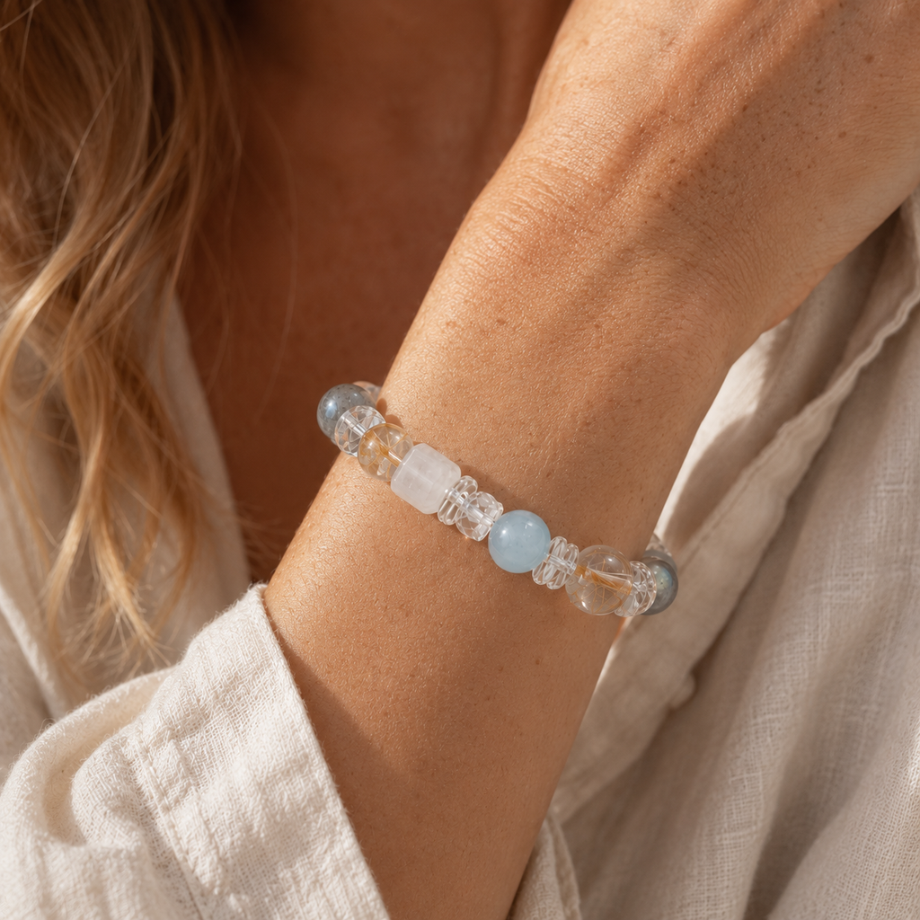 Close-up of a European woman wearing a handmade natural grey moonstone bracelet featuring 10mm aquamarine, golden rutilated quartz, white quartz cube beads, clear quartz cloud spacers and faceted clear quartz accents, styled with cream linen clothing in soft natural window light.