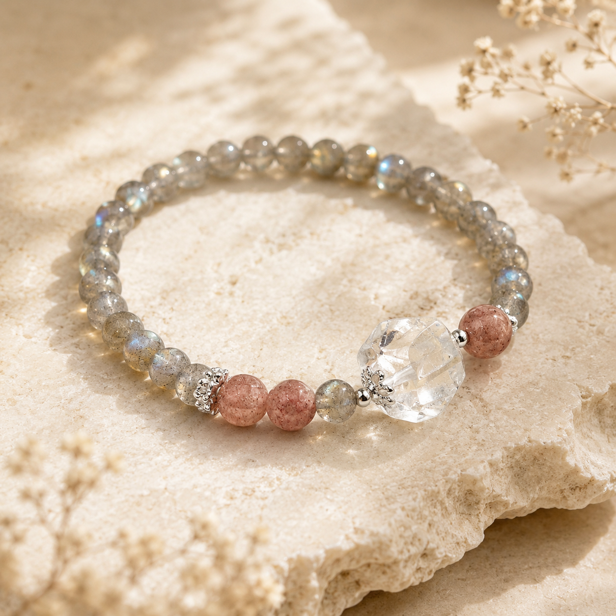 Lifestyle image of a handmade natural grey moonstone bracelet featuring 4mm grey moonstone beads, a freeform clear quartz focal bead, strawberry quartz accent beads and silver-tone spacer beads, displayed on a natural stone surface in soft sunlight.