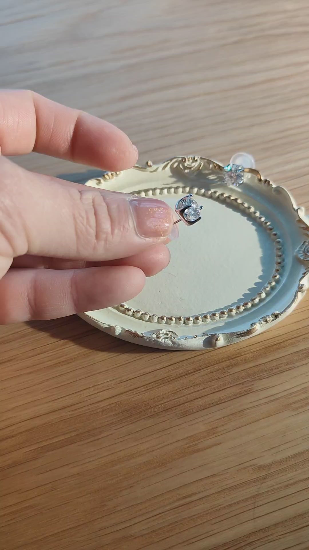 Close-up of a delicate silver moissanite stud earring held between fingers above a decorative jewellery tray in natural sunlight.