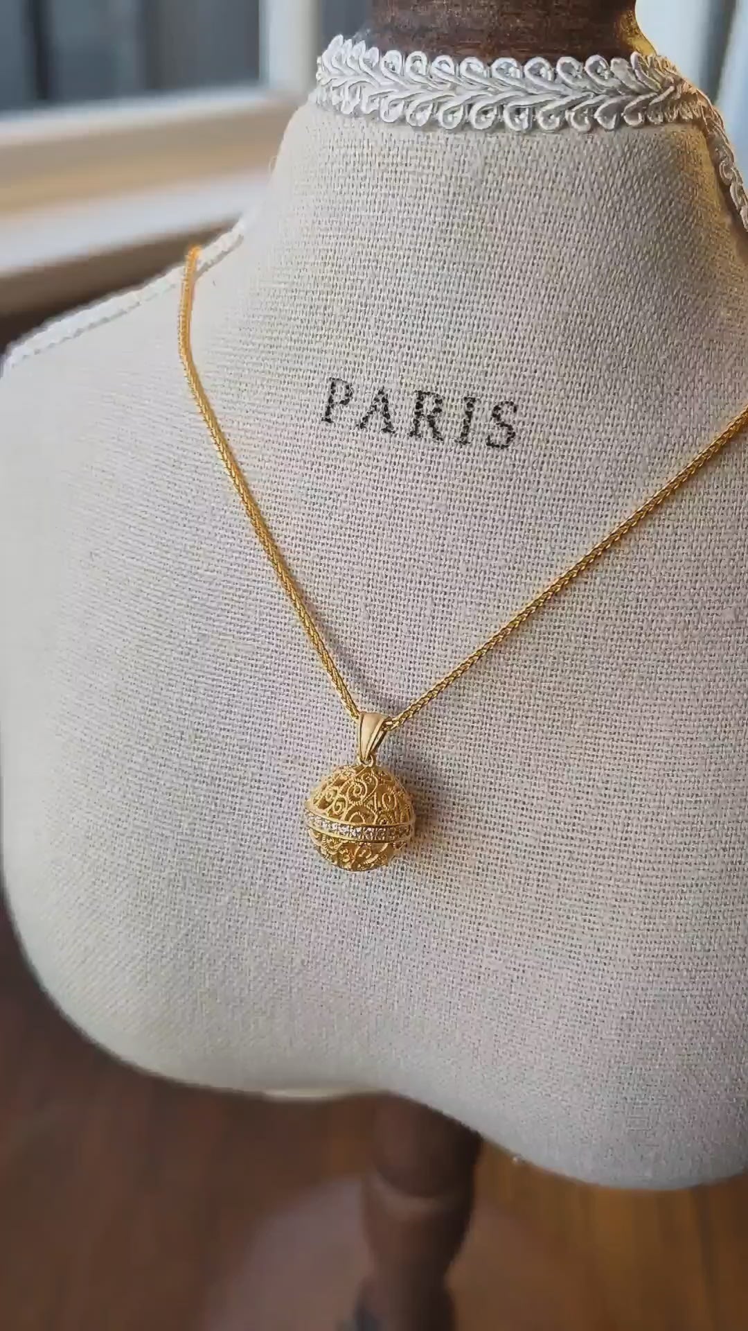 Delicate gold necklace featuring a detailed spherical pendant, photographed on a vintage linen jewellery display in warm natural light.