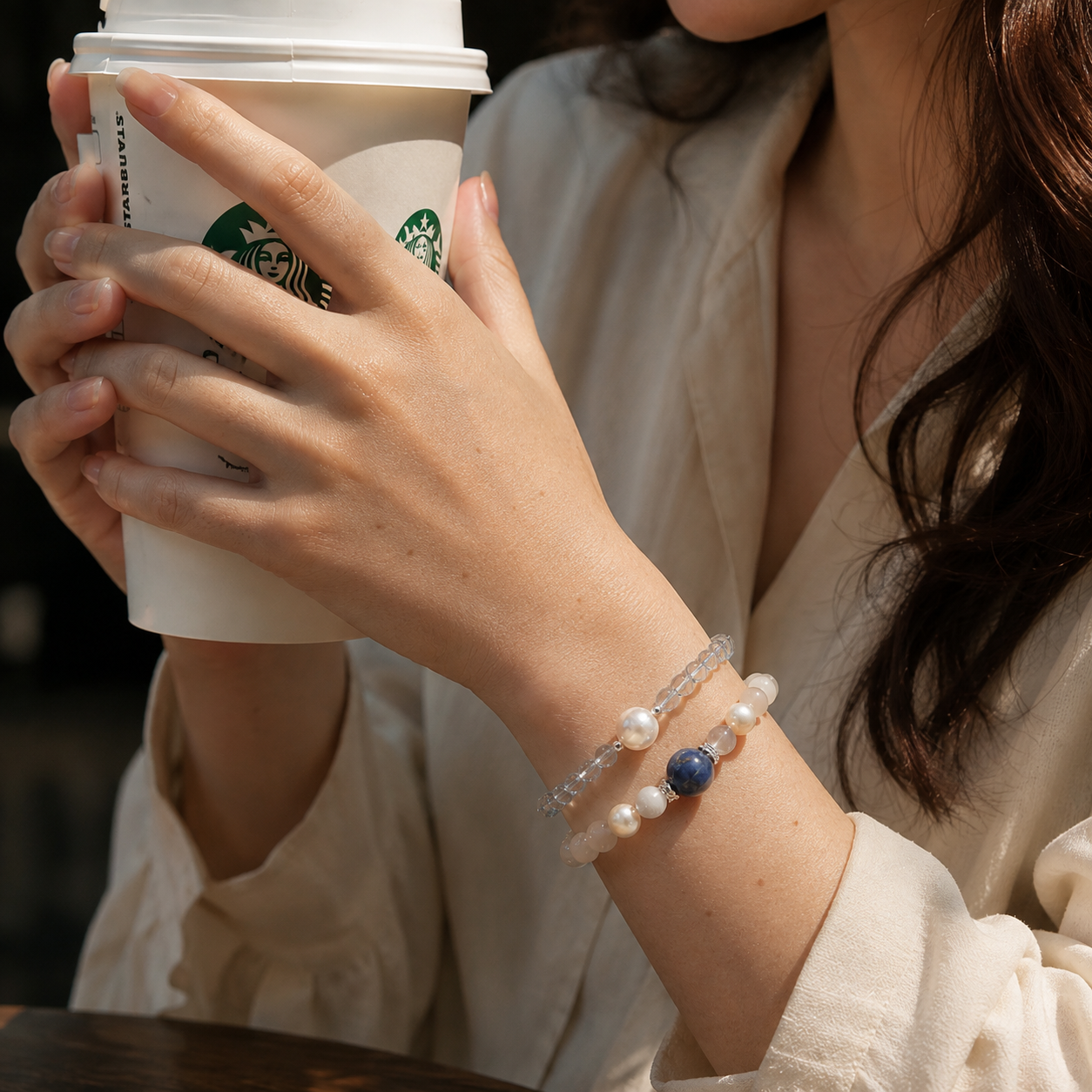 Close-up lifestyle photo of a woman holding a takeaway coffee while wearing stacked handmade gemstone bracelets featuring aquamarine, freshwater pearls, moonstone and a blue natural stone. Elegant everyday jewelry styled for a casual café setting.